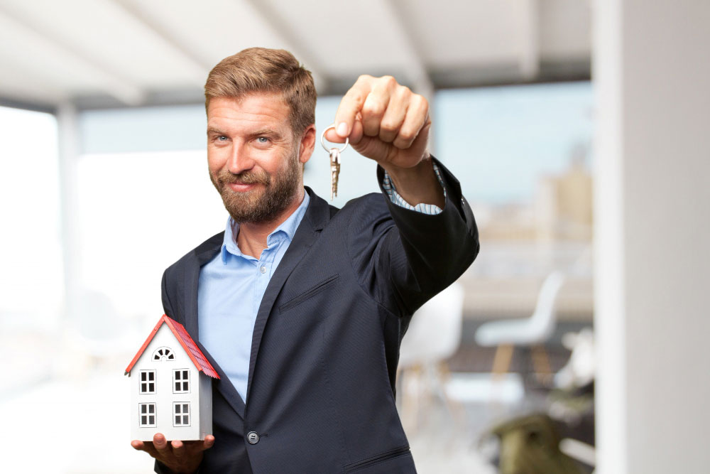 Muž stojící v popředí, v jedné ruce drží miniaturu domečku a ve druhé klíče od domu.