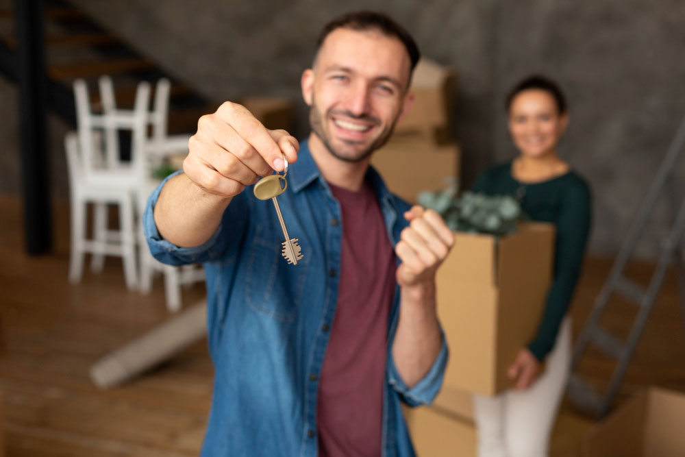 Muž stojící v popředí drží klíče od domu a v pozadí je žena, která drží krabici plnou věcí.